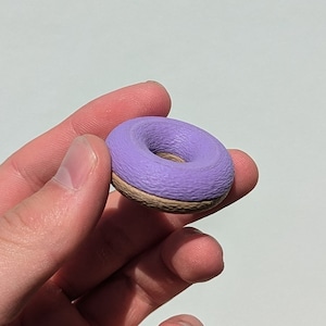 Könnte beinhalten: Ein kleiner lilafarbener Donut mit braunem Kern. Der Donut wird in einer Hand gehalten.