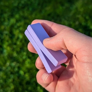 Könnte beinhalten: Ein lilafarbener Plastikclip, der in der Mitte gefaltet ist und in einer Hand gehalten wird.