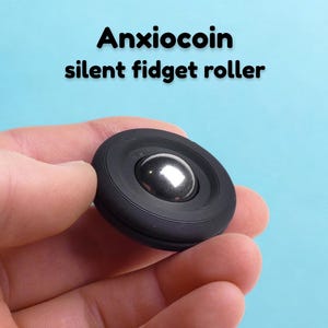 Könnte beinhalten: Ein schwarzer, kreisförmiger Anxiocoin Silent Fidget Roller mit einer glatten, metallisch silbernen Kugel in der Mitte. Der äußere Rand des Geräts weist eine texturierte, geriffelte Oberfläche auf. Der Text Anxiocoin silent fidget roller steht darüber.