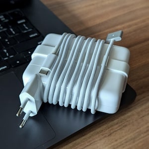 Könnte beinhalten: Ein weißes Laptop-Ladegerät mit einem grauen geflochtenen Kabel, das auf einem schwarzen Laptop liegt. Das Ladegerät ist an einen Netzadapter angeschlossen. Der Laptop befindet sich auf einer Holzoberfläche.
