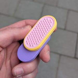 May include: A pair of colourful, ergonomic nail files. The top file is pink with a yellow border and a textured surface. The bottom file is purple. The files are held in a hand against a grey background.