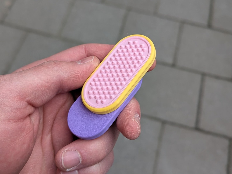 May include: A pair of colourful, ergonomic nail files. The top file is pink with a yellow border and a textured surface. The bottom file is purple. The files are held in a hand against a grey background.