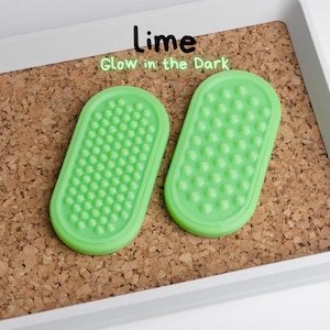 May include: Two lime green, oval-shaped items with textured surfaces, labelled "Lime Glow in the Dark." The items are set on a corkboard within a white tray. The textured surfaces are covered in small, rounded bumps.