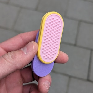 May include: A close-up of a colourful flip-flop. The flip-flop has a purple sole, a pink footbed with small bumps, and a yellow border. The flip-flop is held in a hand.