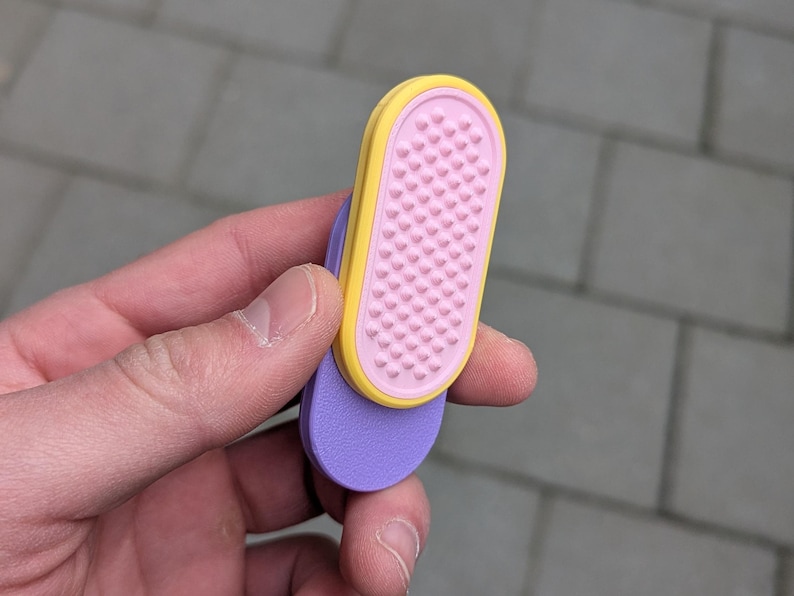 May include: A close-up of a colourful flip-flop. The flip-flop has a purple sole, a pink footbed with small bumps, and a yellow border. The flip-flop is held in a hand.