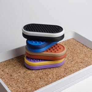 May include: A stack of multi-coloured, oval silicone pads with textured surfaces. The pads are black, blue, orange, yellow, and purple, stacked on a cork tray with a white border. The background is plain white.
