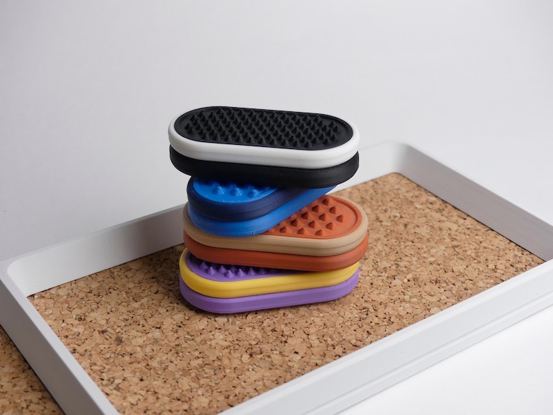 May include: A stack of multi-coloured, oval silicone pads with textured surfaces. The pads are black, blue, orange, yellow, and purple, stacked on a cork tray with a white border. The background is plain white.