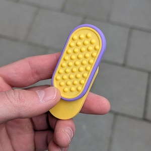 May include: A yellow and purple phone grip with a textured surface. The grip is held in a hand and has a rectangular shape with rounded edges. The grip is designed to attach to the back of a phone.