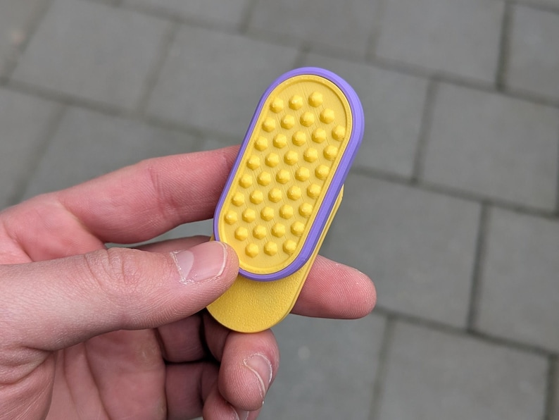 May include: A yellow and purple phone grip with a textured surface. The grip is held in a hand and has a rectangular shape with rounded edges. The grip is designed to attach to the back of a phone.