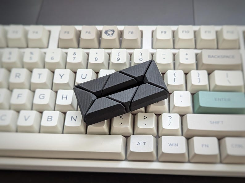 May include: A black plastic keyboard spacer with a geometric design. The spacer is placed on a white keyboard with black keys.