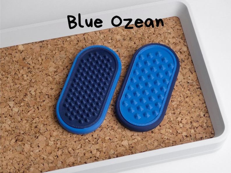 May include: Two blue oval pet grooming tools with textured surfaces are displayed on a corkboard. The text "Blue Ozean" is written in black above the objects. The tools are in a white tray.