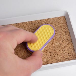 May include: A yellow and purple oval-shaped object with a textured surface is held over a corkboard. The object is inside a white rectangular tray. The corkboard is a light brown colour.