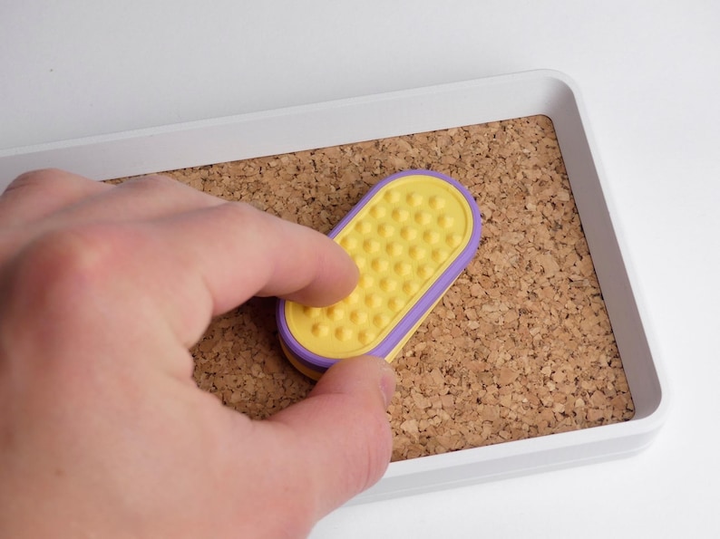 May include: A yellow and purple oval-shaped object with a textured surface is held over a corkboard. The object is inside a white rectangular tray. The corkboard is a light brown colour.