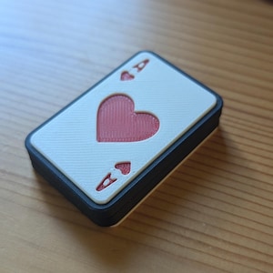 May include: A white playing card with a black border, featuring a red heart and the ace of hearts symbol. The card is made of a textured material.