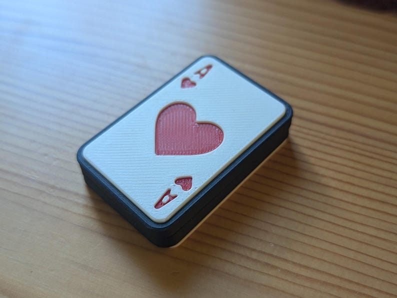 May include: A white playing card with a black border, featuring a red heart and the ace of hearts symbol. The card is made of a textured material.