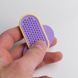 May include: A purple and yellow nail file with a textured surface. The nail file is held in a hand against a white background. The file has a rounded rectangular shape with a yellow border.