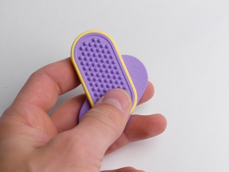 May include: A purple and yellow nail file with a textured surface. The nail file is held in a hand against a white background. The file has a rounded rectangular shape with a yellow border.