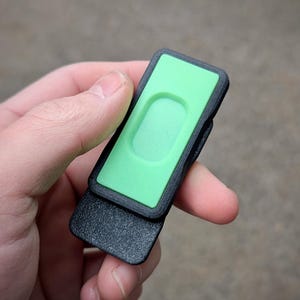 Op de afbeelding: Een zwarte en groene plastic clip met een afgeronde knop. De clip is ontworpen om aan een riem of tas te worden bevestigd.