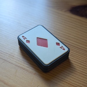 May include: A white playing card with a red diamond suit and the number 'A' printed on it. The card is framed with a black border and is sitting on a wooden surface.