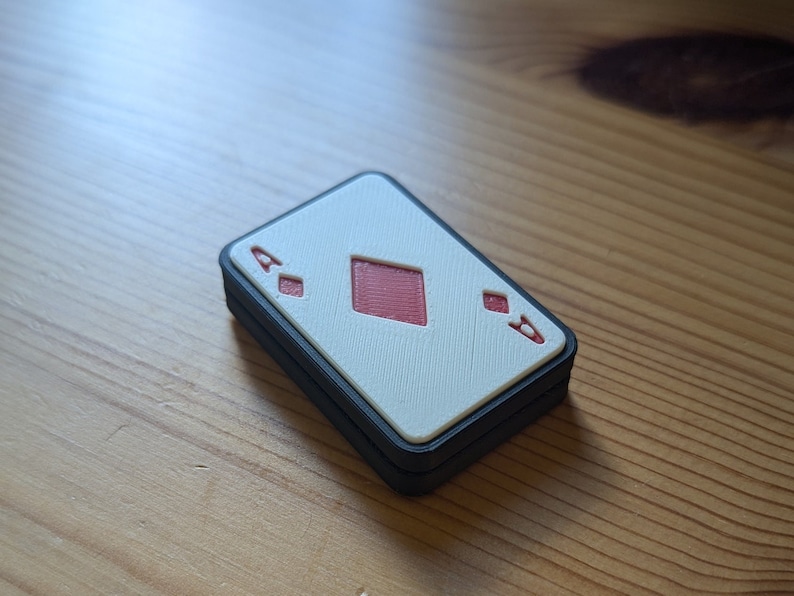 May include: A white playing card with a red diamond suit and the number 'A' printed on it. The card is framed with a black border and is sitting on a wooden surface.