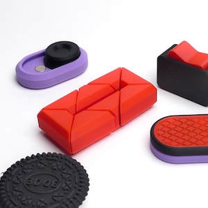 May include: Five different silicone objects in various colors and shapes. A red object with a geometric design, a purple object with a black circle, a black object with a red button, a purple object with a red and black patterned surface, and a black cookie with the word "EDGE" printed on it.