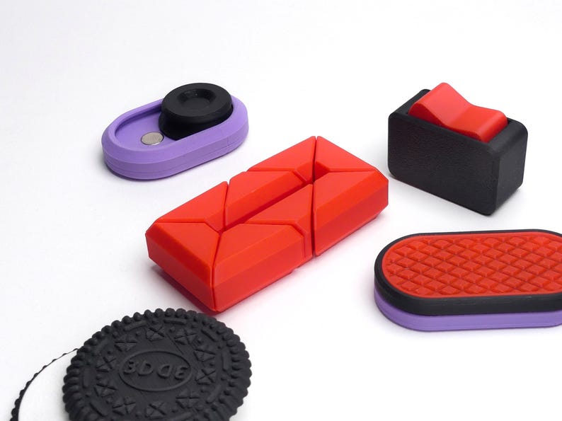May include: Five different silicone objects in various colors and shapes. A red object with a geometric design, a purple object with a black circle, a black object with a red button, a purple object with a red and black patterned surface, and a black cookie with the word "EDGE" printed on it.
