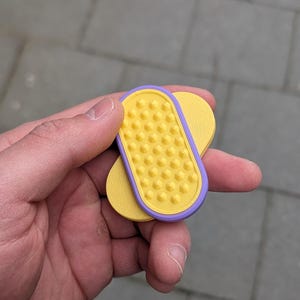 May include: A yellow and purple pet grooming tool with a textured surface. The tool is held in a hand and appears to be made of plastic. The tool has a rectangular shape with rounded edges and a series of small, raised bumps.