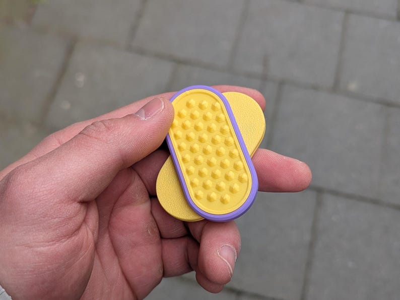 May include: A yellow and purple pet grooming tool with a textured surface. The tool is held in a hand and appears to be made of plastic. The tool has a rectangular shape with rounded edges and a series of small, raised bumps.