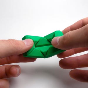 May include: A green, flexible, geometric puzzle toy made of interlocking triangular pieces. The toy is being held in two hands, demonstrating its ability to bend and fold.