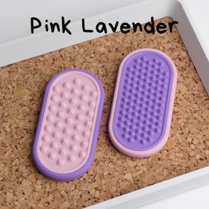 May include: Two oval-shaped, textured items in pink and purple, labelled "Pink Lavender". The items are set on a corkboard surface within a white tray. One item is pink with pink bumps, the other is purple with purple bumps.