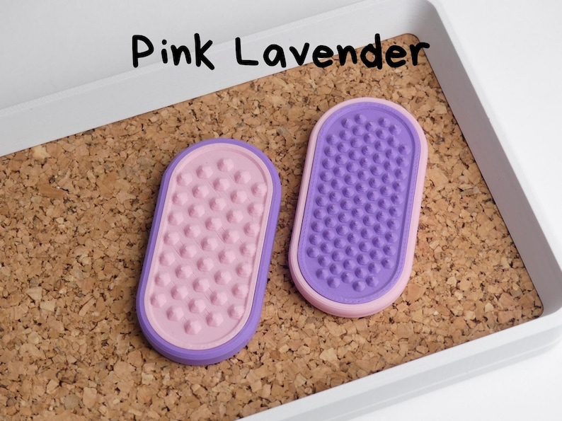 May include: Two oval-shaped, textured items in pink and purple, labelled "Pink Lavender". The items are set on a corkboard surface within a white tray. One item is pink with pink bumps, the other is purple with purple bumps.