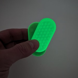 May include: A bright green, handheld device with a textured surface and rounded edges. The device is held in a hand against a neutral background. The device appears to be made of a flexible material.