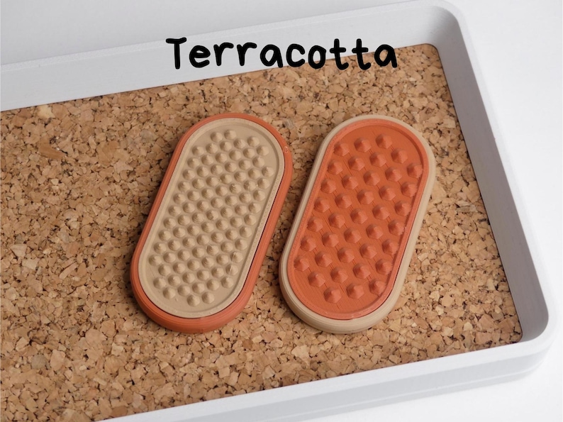 May include: Two terracotta-coloured, oval-shaped objects with textured surfaces, displayed on a corkboard within a white tray. The word "Terracotta" is written in black above the objects. The objects have a beige and orange colour scheme.