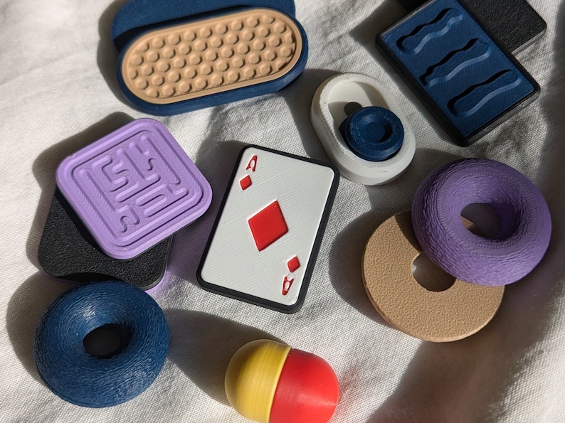 May include: A collection of colorful, textured, and patterned objects, including a playing card with the Ace of Diamonds, a purple donut, a blue donut, a purple square with a pattern, a brown circle with a hole in the center, a blue and white object with a wavy pattern, a yellow and red pill-shaped object, and a white object with a blue circle.