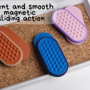 May include: Three magnetic sliding erasers in orange, blue, and pink, with a textured surface. The erasers are displayed on a corkboard within a white tray. The text "silent and smooth magnetic sliding action" is visible.