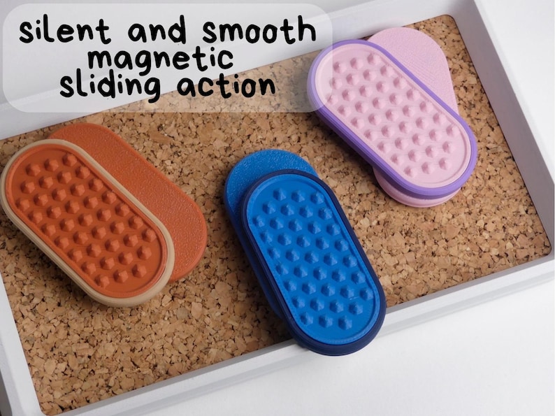 May include: Three magnetic sliding erasers in orange, blue, and pink, with a textured surface. The erasers are displayed on a corkboard within a white tray. The text "silent and smooth magnetic sliding action" is visible.