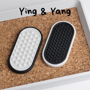 May include: Two oval coasters, one white with a black border and white dots, the other black with a white border and a textured surface. The coasters are on a cork surface inside a white tray. The text "Ying & Yang" is above the coasters.