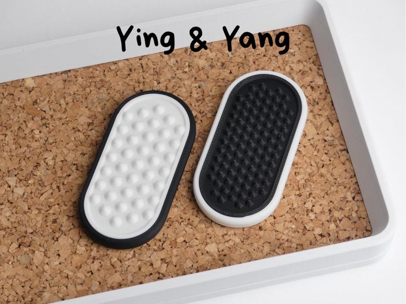 May include: Two oval coasters, one white with a black border and white dots, the other black with a white border and a textured surface. The coasters are on a cork surface inside a white tray. The text "Ying & Yang" is above the coasters.