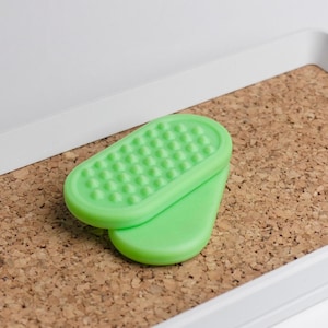 May include: Two-piece green foot care product. The top piece has a textured surface with small, rounded bumps. The bottom piece is smooth. The items are on a cork surface, inside a white tray.