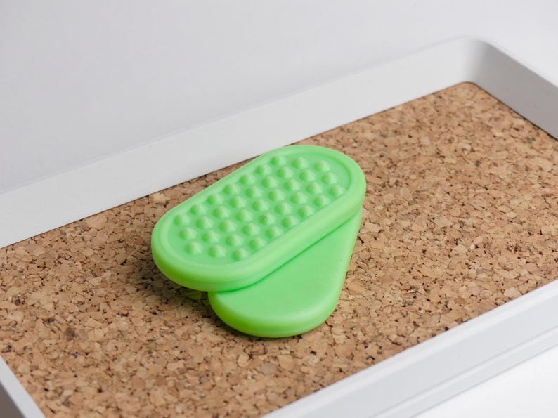 May include: Two-piece green foot care product. The top piece has a textured surface with small, rounded bumps. The bottom piece is smooth. The items are on a cork surface, inside a white tray.
