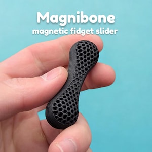 Pode incluir: Magnibone magnético preto com design de favo de mel. O texto "Magnibone magnetic fidget slider" é branco. O brinquedo fidget é segurado contra um fundo azul claro.