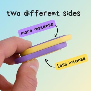 May include: A yellow and purple massage tool with two different sides, held in a hand. The image text reads "two different sides", "more intense", and "less intense". The tool is designed for massage.