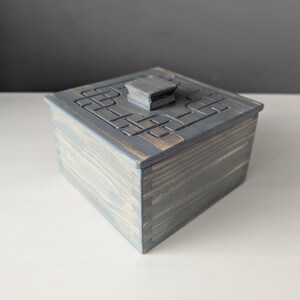 May include: A small, square wooden box with a hinged lid. The box is painted a dark grey colour and has a carved design on the top. The lid has a small knob on top.