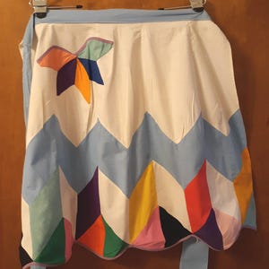 May include: A colorful, handmade apron with a white base and a blue top and bottom. The apron features a zig-zag pattern in various colors, including orange, purple, and green. A star-shaped applique is on the upper part of the apron.