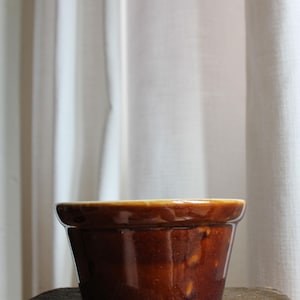 May include: A brown ceramic crock pot with a glossy finish. The pot has a wide, slightly flared rim and a rounded base. It sits on a weathered wooden surface, with a white curtain in the background.