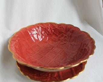 May include: A red ceramic serving bowl with a floral design and gold trim. The bowl has a diamond pattern and a scalloped edge. The bowl is likely used for serving food or as a decorative piece.