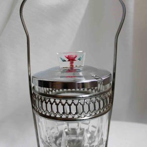 May include: A vintage sugar bowl with a clear glass body and a decorative silver-tone metal frame and handle. The lid features a small, square glass knob with a red and green floral design. The bowl is designed to hold sugar or other condiments.
