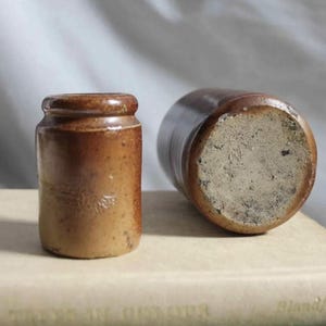 Peut inclure: Deux pots anciens en grès brun. L'un est droit, cylindrique avec un petit rebord. L'autre est couché, révélant une base texturée. Les pots sont posés sur une surface de couleur claire.