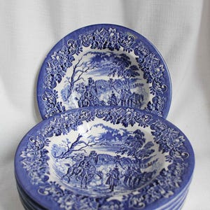 May include: A stack of blue and white ceramic bowls. The bowls feature a detailed pastoral scene and floral border. The bowls are stacked on top of each other, with the top bowl showing the full design. The bowls are likely for serving.