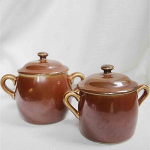 May include: Two small, brown ceramic pots with lids. Each pot has a rounded body, two small handles, and a matching lid with a small knob. The pots are a warm, earthy brown colour and appear to be glazed.